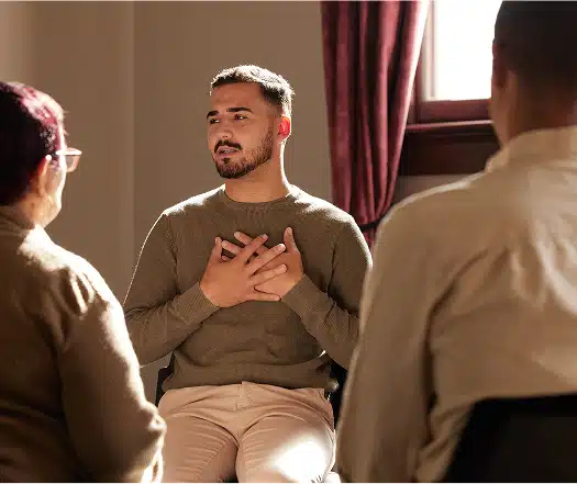 A man sitting in a chair with his hands over his heart expresses himself during a therapy session.