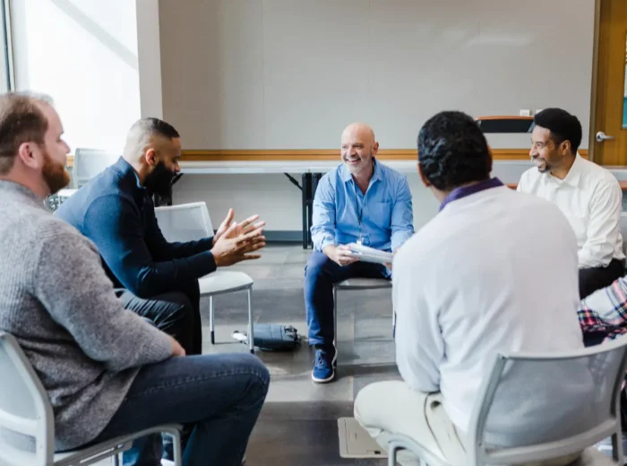 Six men sit in a circle in a therapy setting as one man talks while others look on smiling.