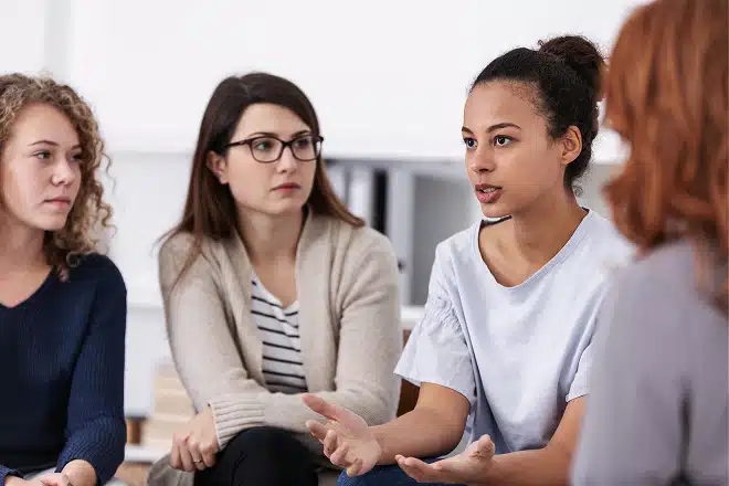 Four women sit together, three looking on as the fourth speaks, during a group therapy session.